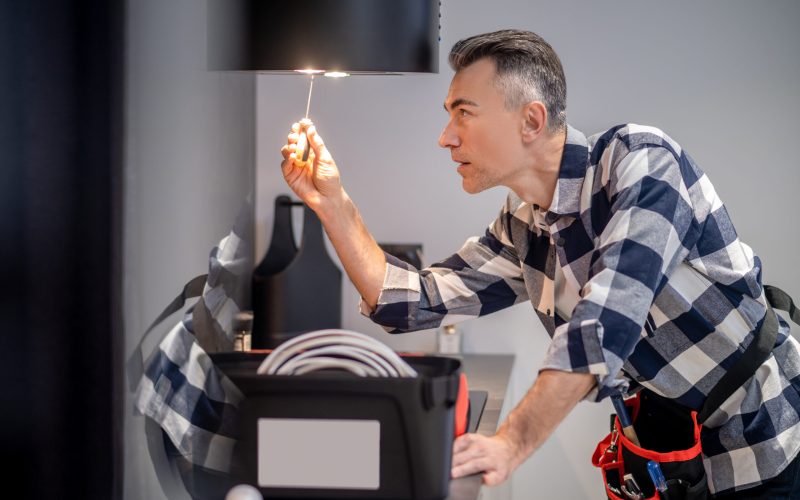 Renovation. Profile of concentrated middle aged man in plaid shirt touching screwdriver to lamp on kitchen range hood in modern kitchen at home